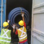 Workers loading a large coil of flexible industrial conduit into a shipping container