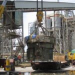 Heavy industrial equipment suspended by crane above a low-bed trailer