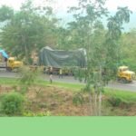Wide view of a heavy transport convoy carrying covered oversized equipment through a rural area