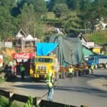 Oversized cargo convoy passing through a roadside community with traffic personnel assisting