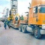 Orange tractor head hauling steel sheet rolls on a low-bed trailer at a wharf